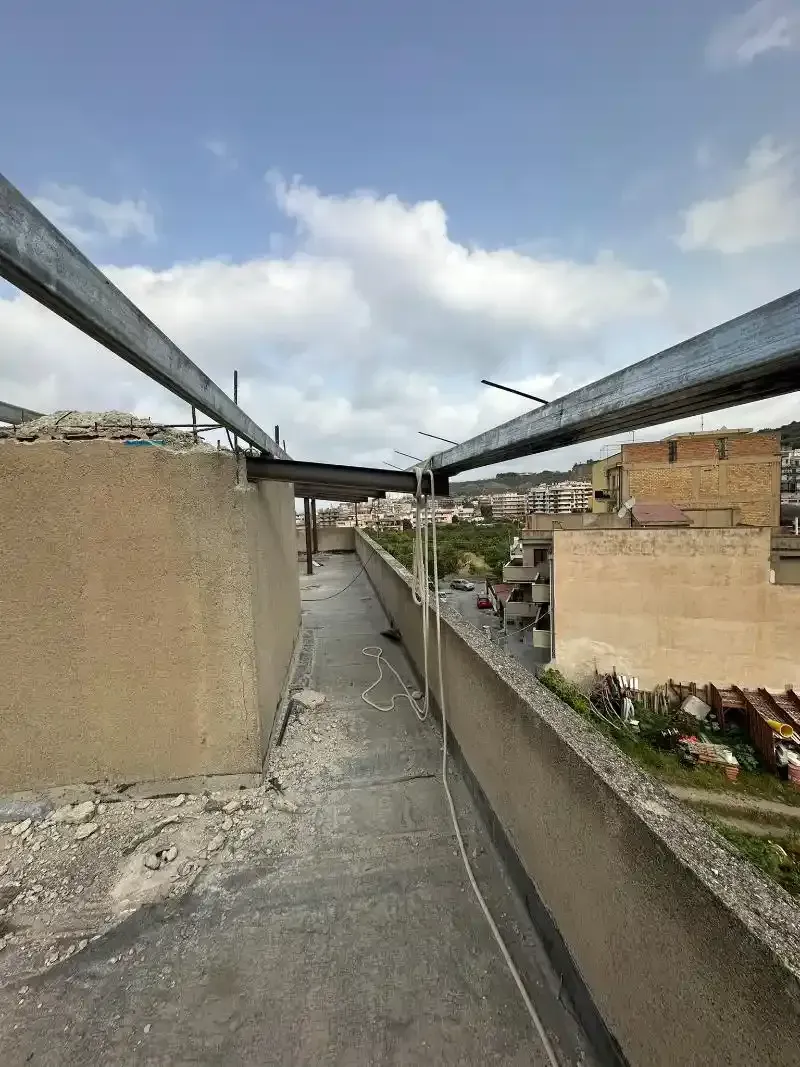 Un tetto con travi metalliche, muri di cemento e un paesaggio urbano in lontananza sotto un cielo nuvoloso.