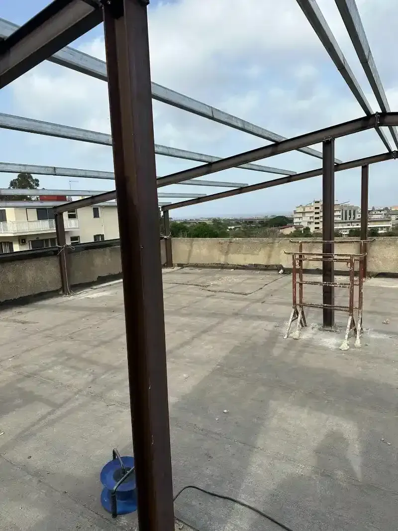 Struttura con telaio in acciaio su un tetto, con una colonna marrone in primo piano e un cielo nuvoloso.