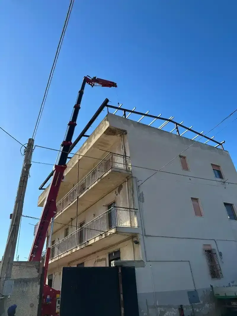 Una gru solleva travi metalliche sul tetto di un edificio di tre piani sotto un cielo azzurro e terso.