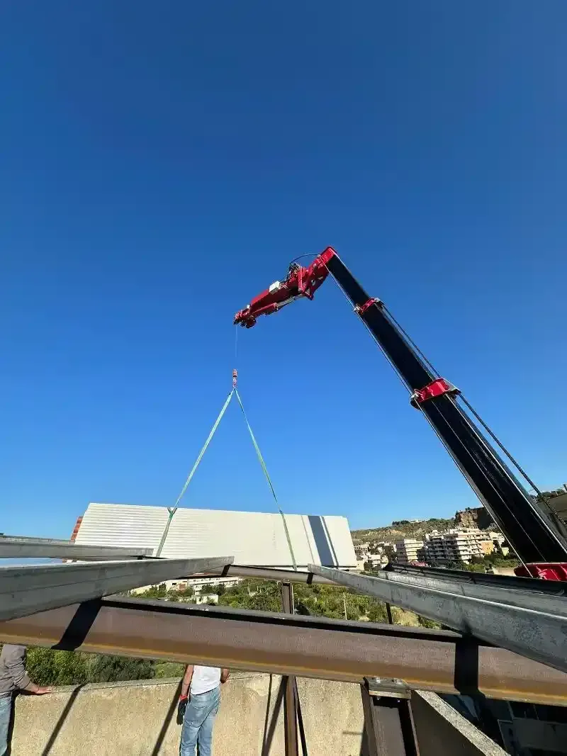 Una gru solleva un grande oggetto rettangolare bianco sulla struttura di un edificio sotto un cielo azzurro.