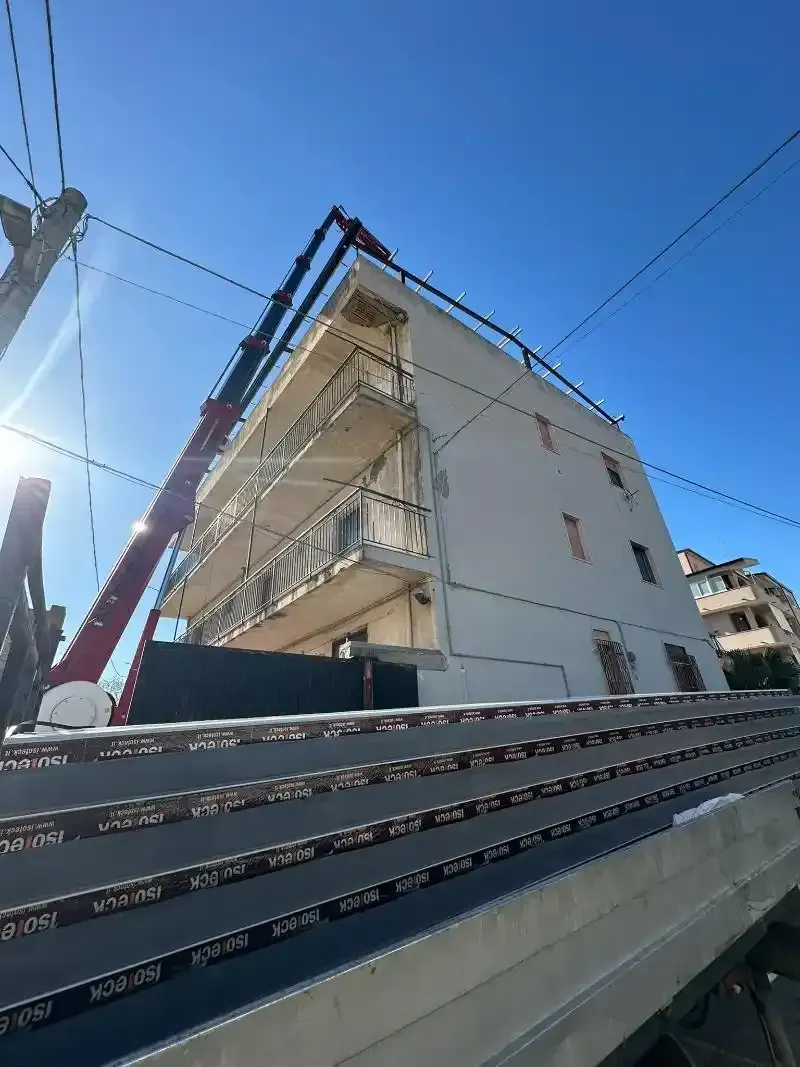Una gru solleva materiali vicino a un edificio con balconi in una giornata di sole.