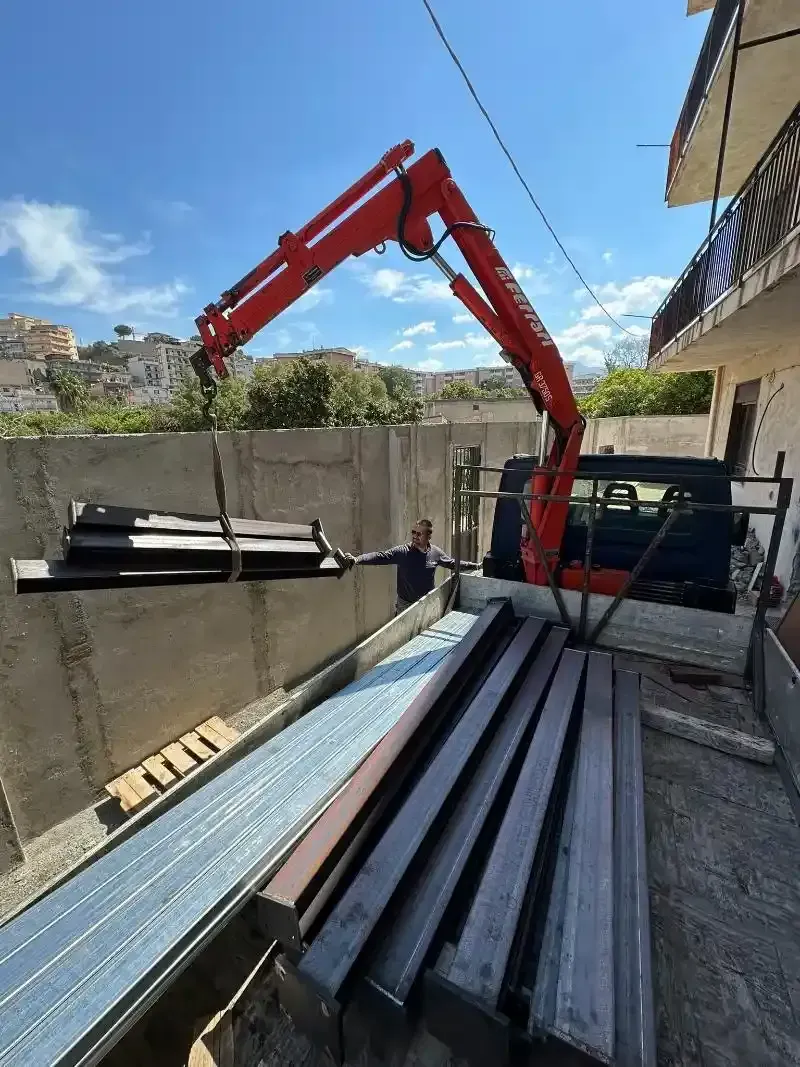 Gru rossa che solleva travi metalliche vicino a un muro di cemento; persona nelle vicinanze; giornata di sole.