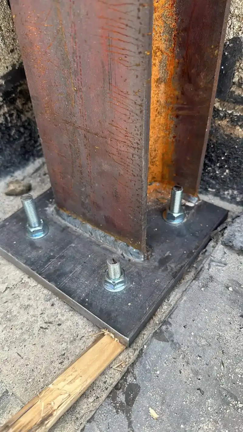 Base in trave d'acciaio imbullonata a una superficie in cemento. Sono visibili segni di ruggine e saldatura.
