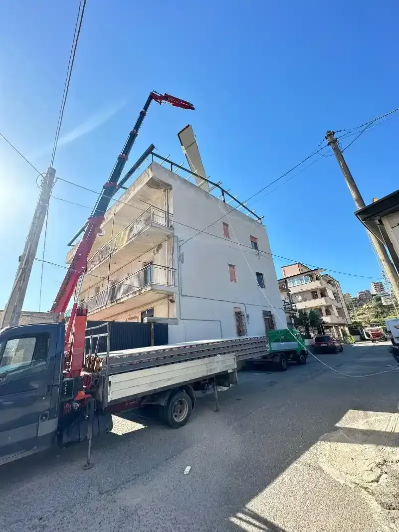 Una gru solleva un grande oggetto bianco sul tetto di un edificio a più piani in una strada soleggiata.