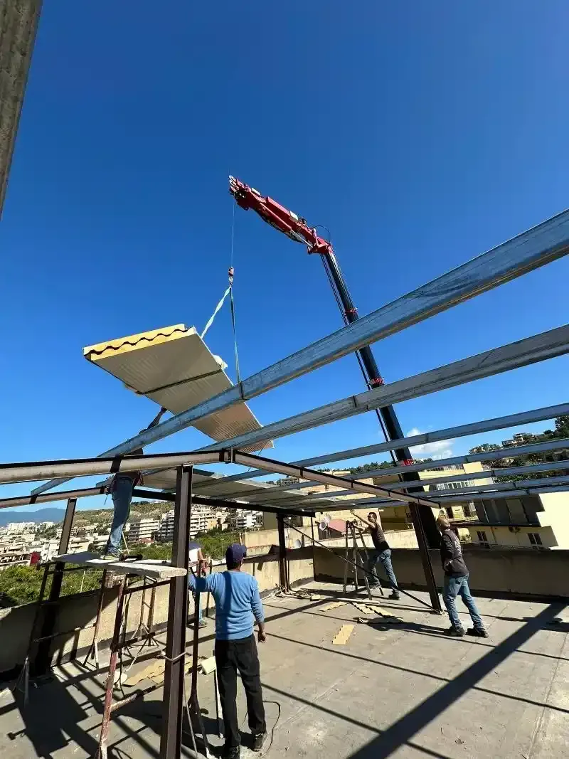 Operai che installano i pannelli del tetto sulla struttura di un edificio con l'aiuto di una gru sotto un cielo azzurro e terso.