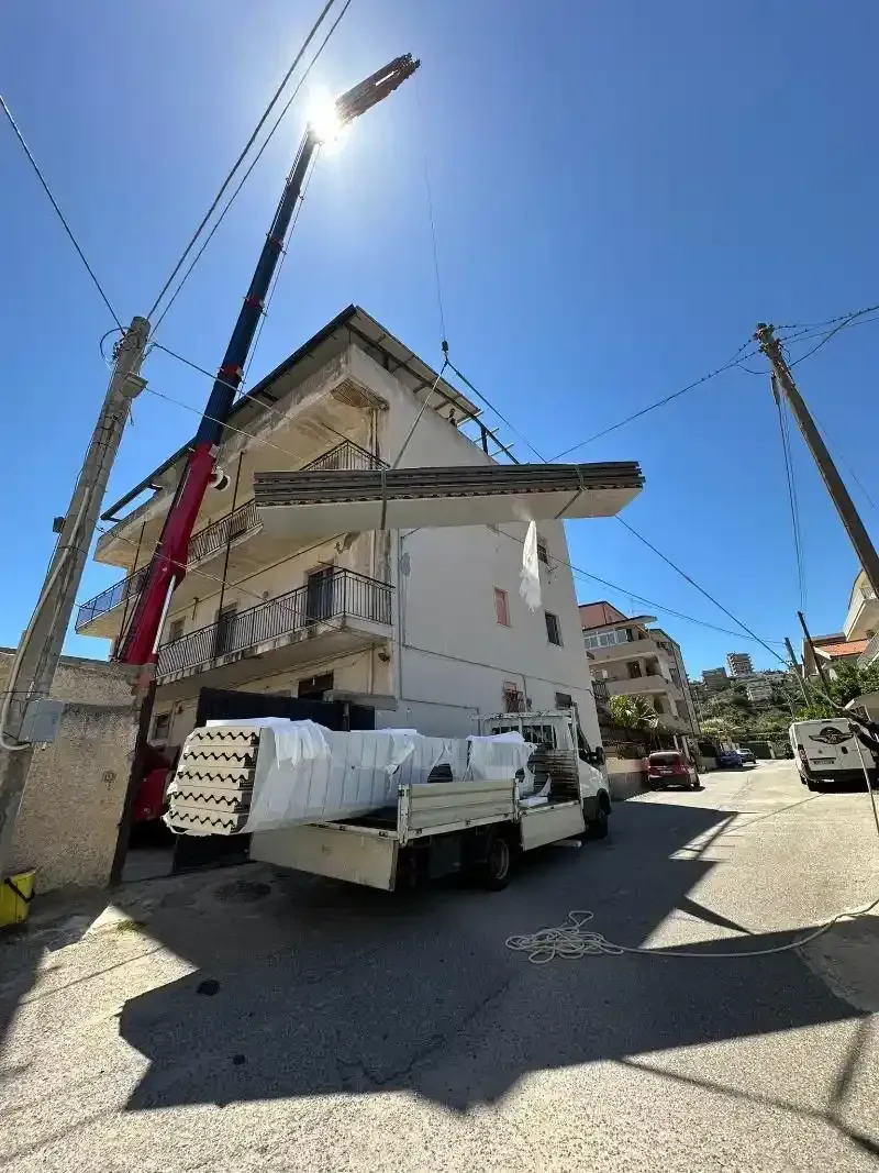 Una gru solleva una lastra di cemento da un edificio di tre piani, passando sopra un camion su una strada soleggiata.