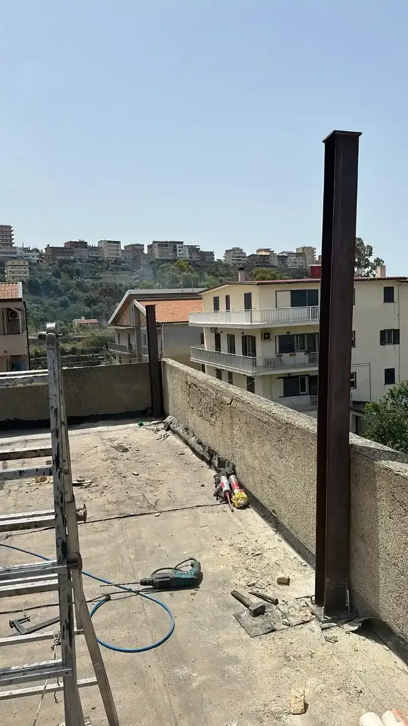 Cantiere con travi metalliche erette su un tetto con vista sugli edifici e su una collina sotto un cielo soleggiato.