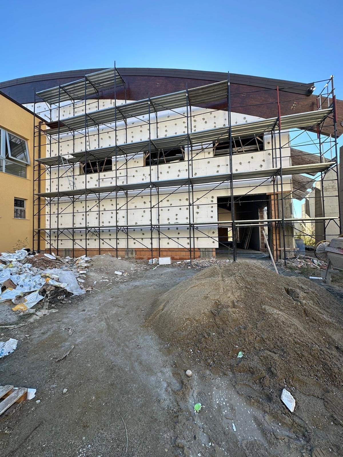 Edificio in costruzione; muri beige, impalcature, reti verdi, cielo azzurro.