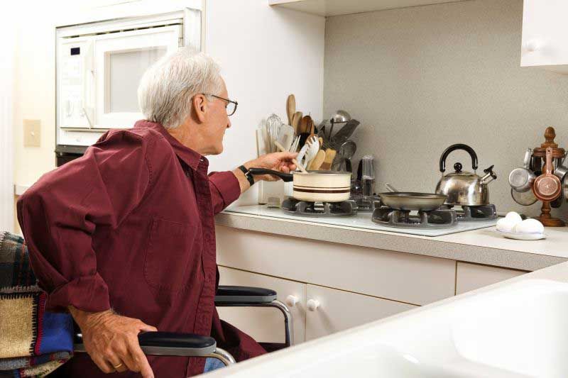 An elderly man in a wheelchair is cooking in the kitchen.