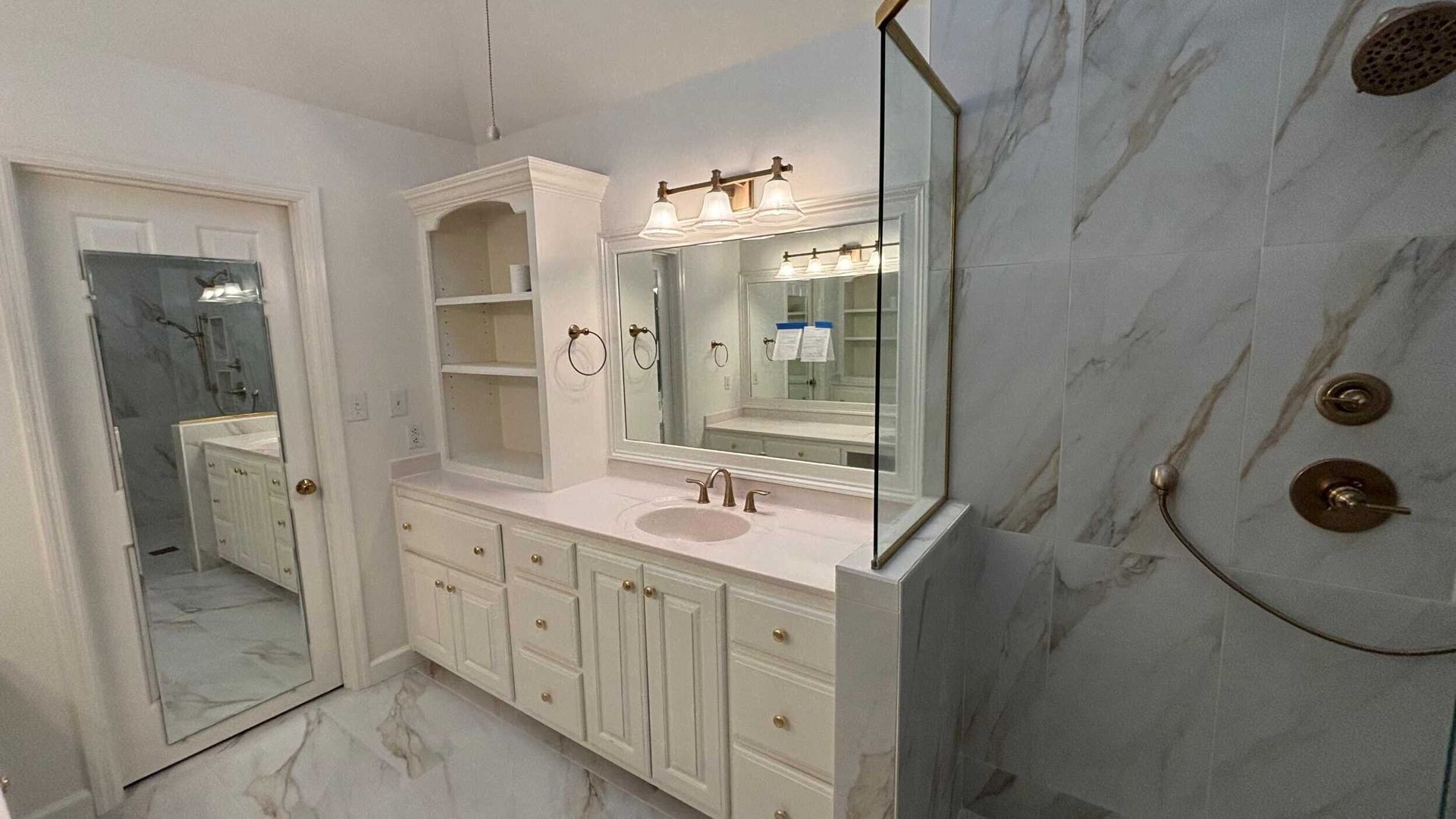 A bright white bathroom with marble tile and a glass shower door. White vanity and mirrored cabinet.