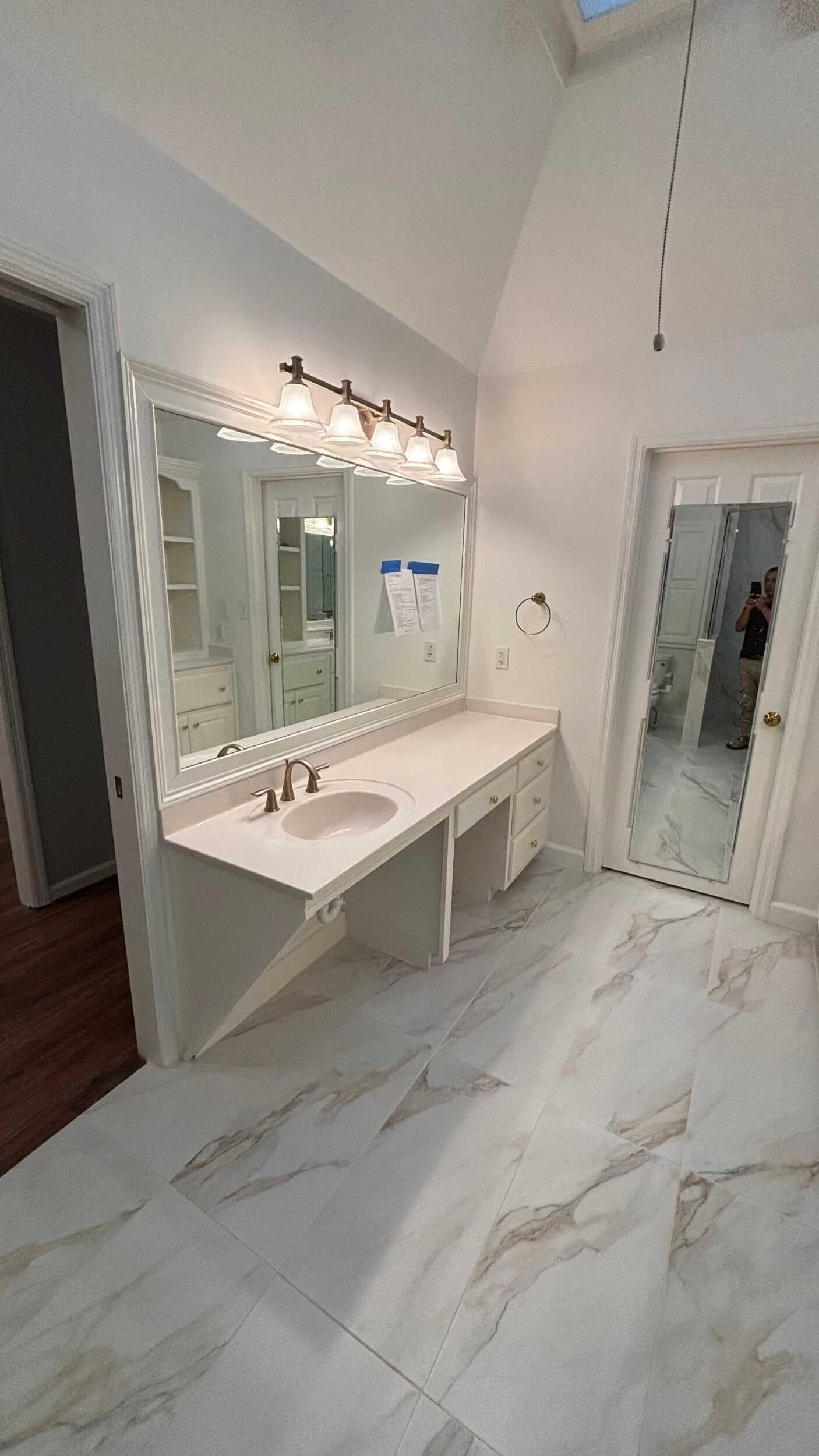 Bathroom with white walls, marble-look floors, long vanity with mirror and lights, and doorway to a separate toilet room.
