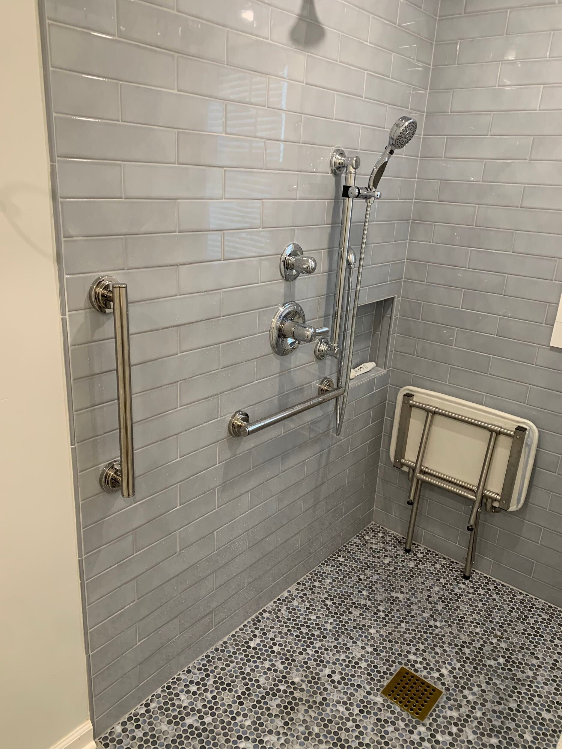 Accessible shower with gray tiled walls, grab bars, fold-down seat, and pebble floor.