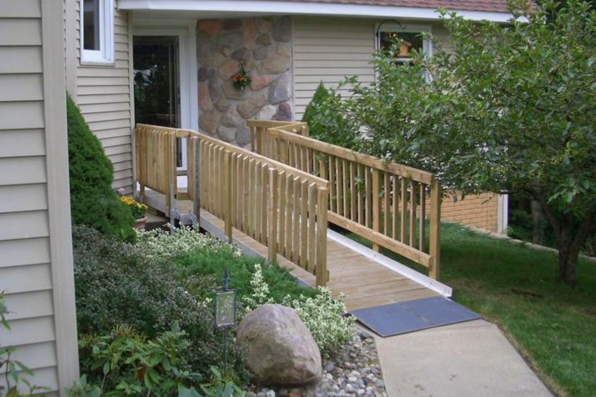 Wooden ramp with handrails leading to a building entrance. Landscaping with shrubs and a tree surround it.