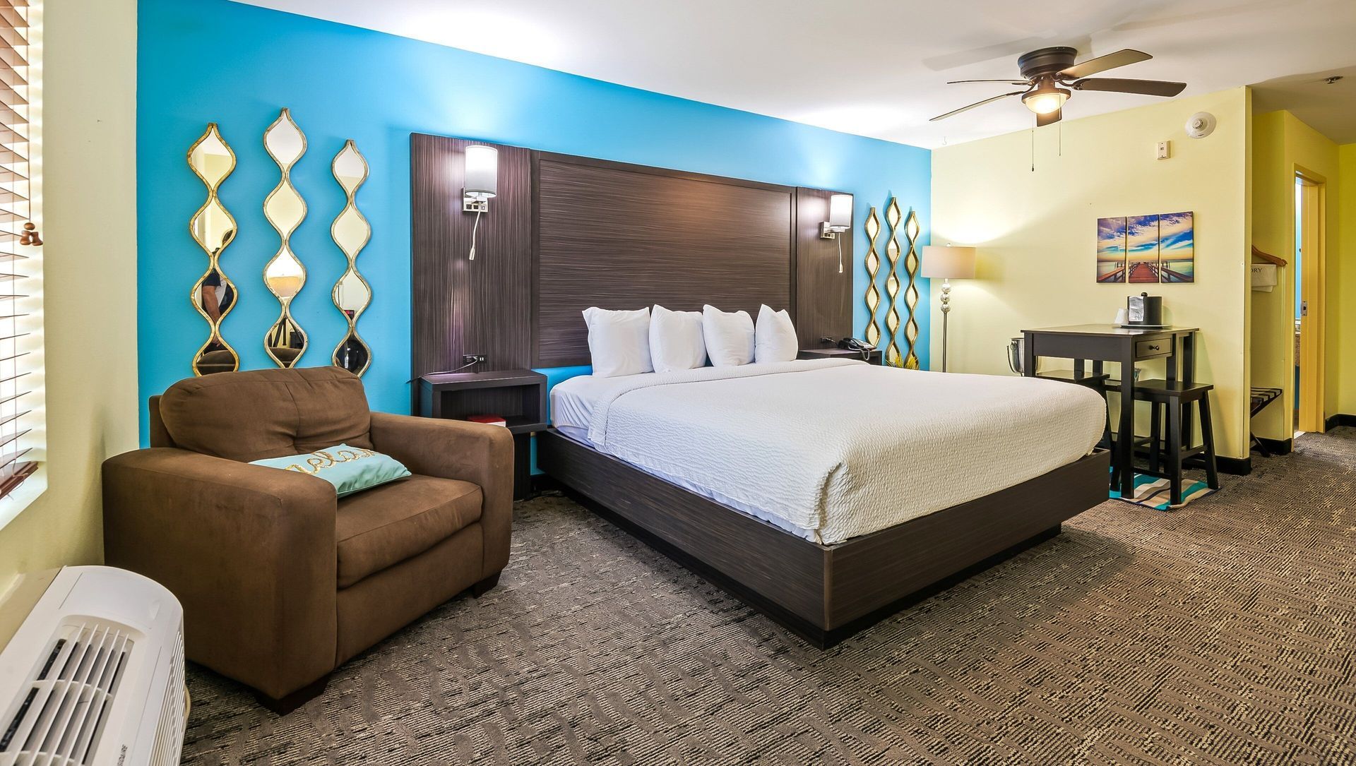 A hotel room with a king size bed , chair , table and ceiling fan.