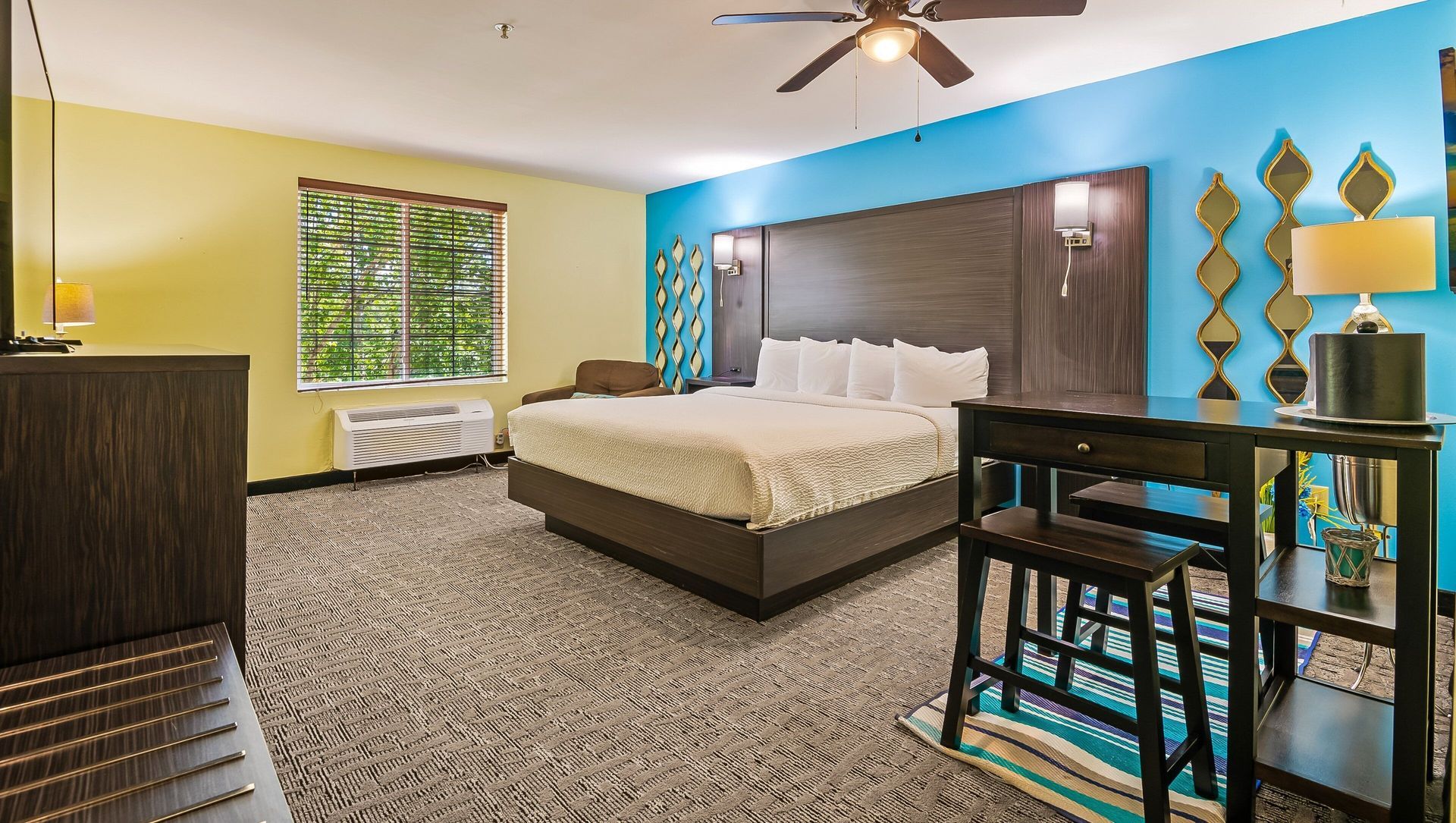 A hotel room with a king size bed , desk , chairs and a ceiling fan.