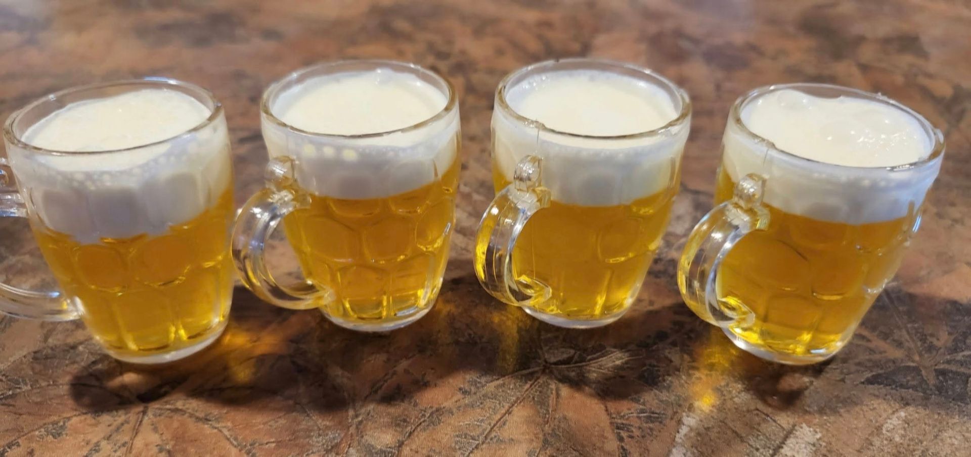 Four small beer mugs filled with beer and foam, on a speckled surface.