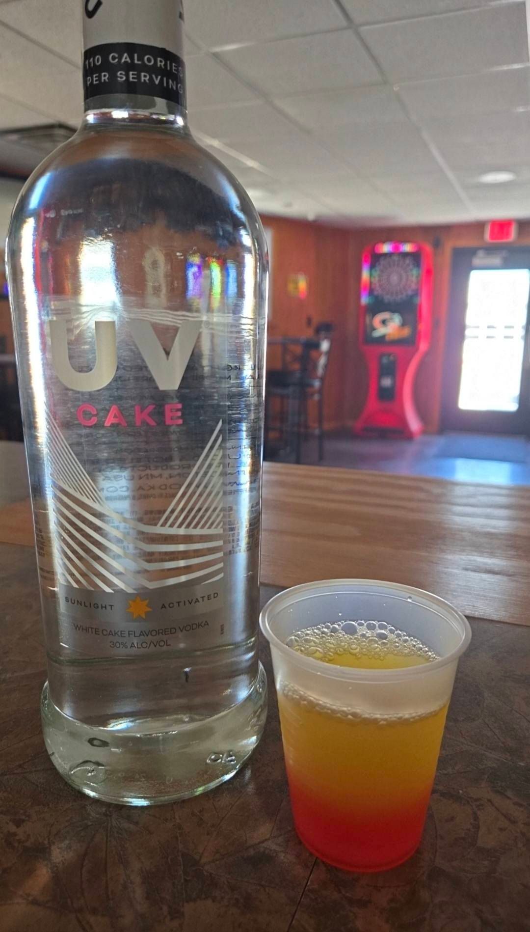 Vodka bottle next to a layered drink in a plastic cup; bar setting with a dartboard.