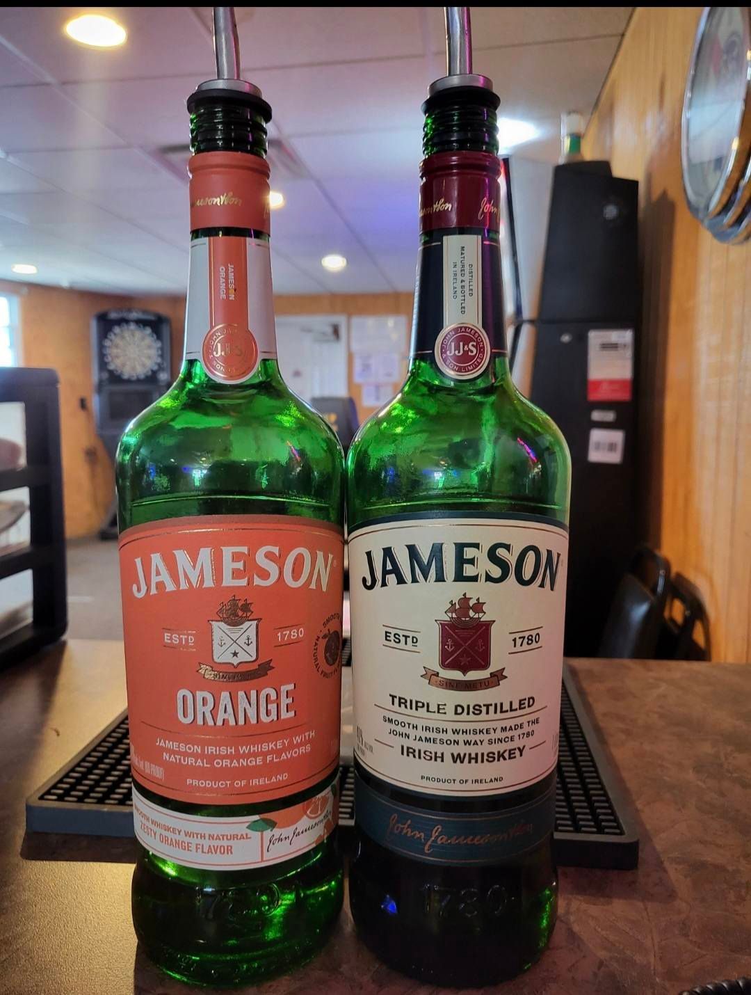 Two Jameson whiskey bottles on a bar, one orange-labeled, the other standard, both with pour spouts.