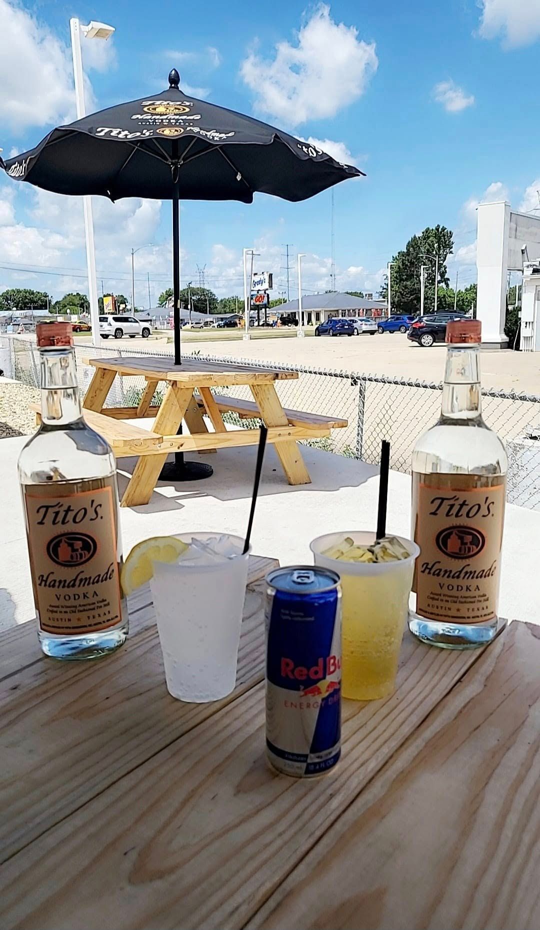Two Tito's bottles, cocktails, Red Bull, wooden picnic table, patio setting with umbrella on a sunny day.