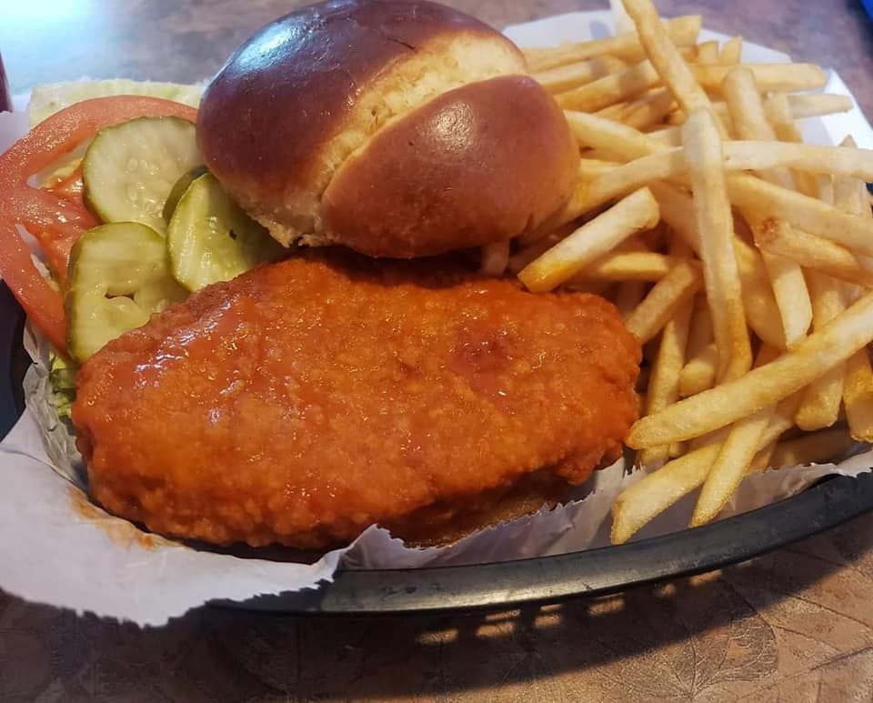 Buffalo chicken sandwich with fries, pickles, tomato, and pretzel bun in a basket.