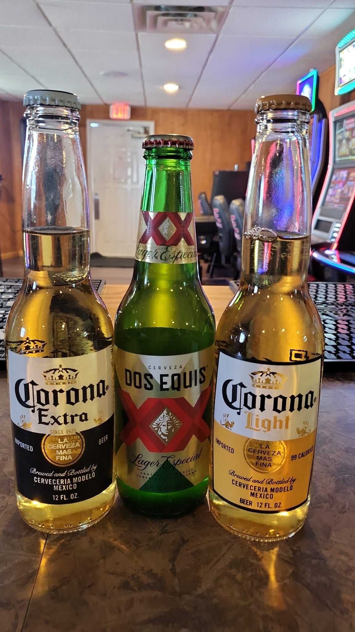 Three beer bottles: Corona Extra, Dos Equis, Corona Light, on a bar counter inside a bar.
