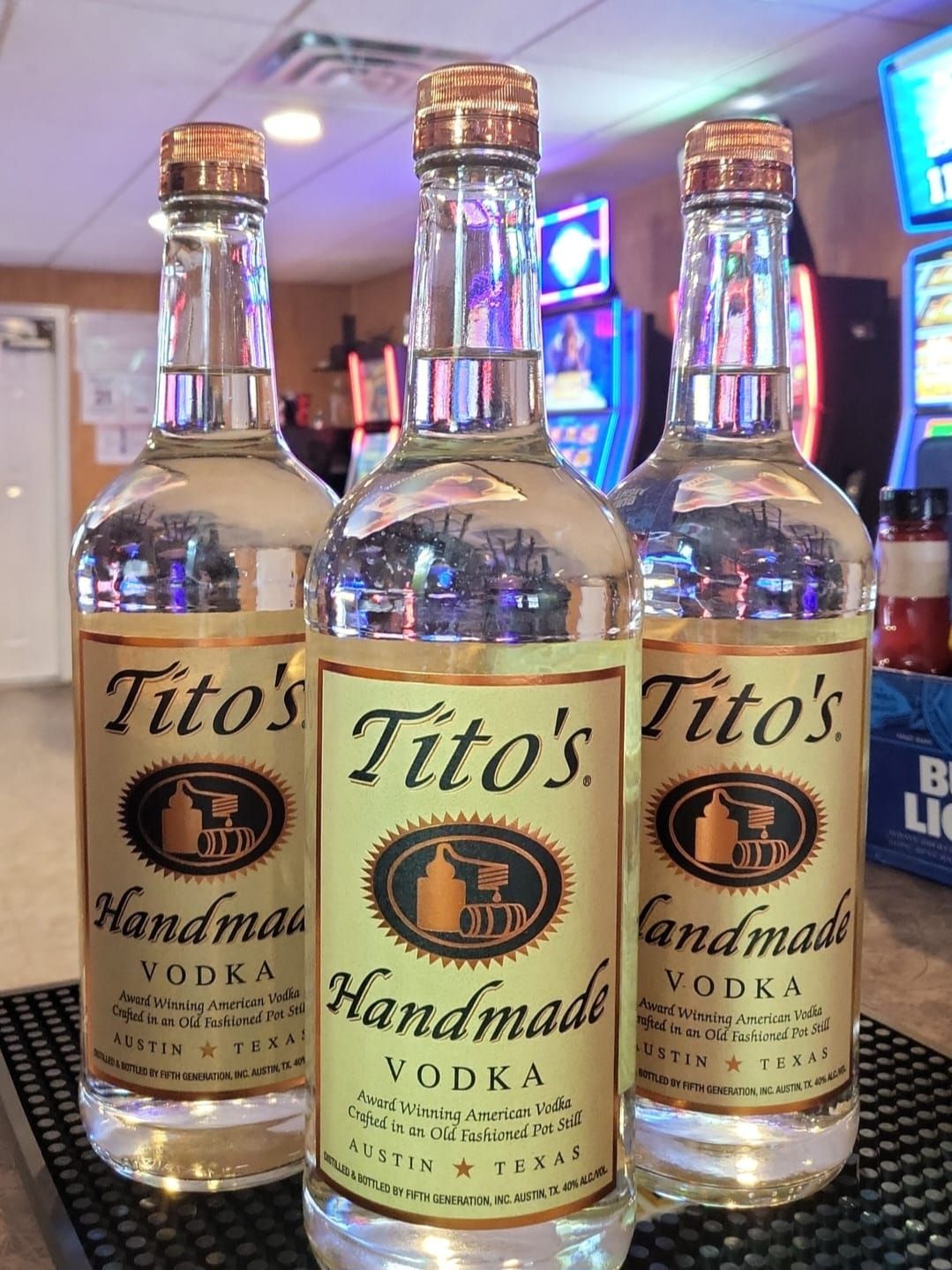 Three bottles of Tito's Handmade Vodka with gold labels, lined up on a bar with slot machines in the background.