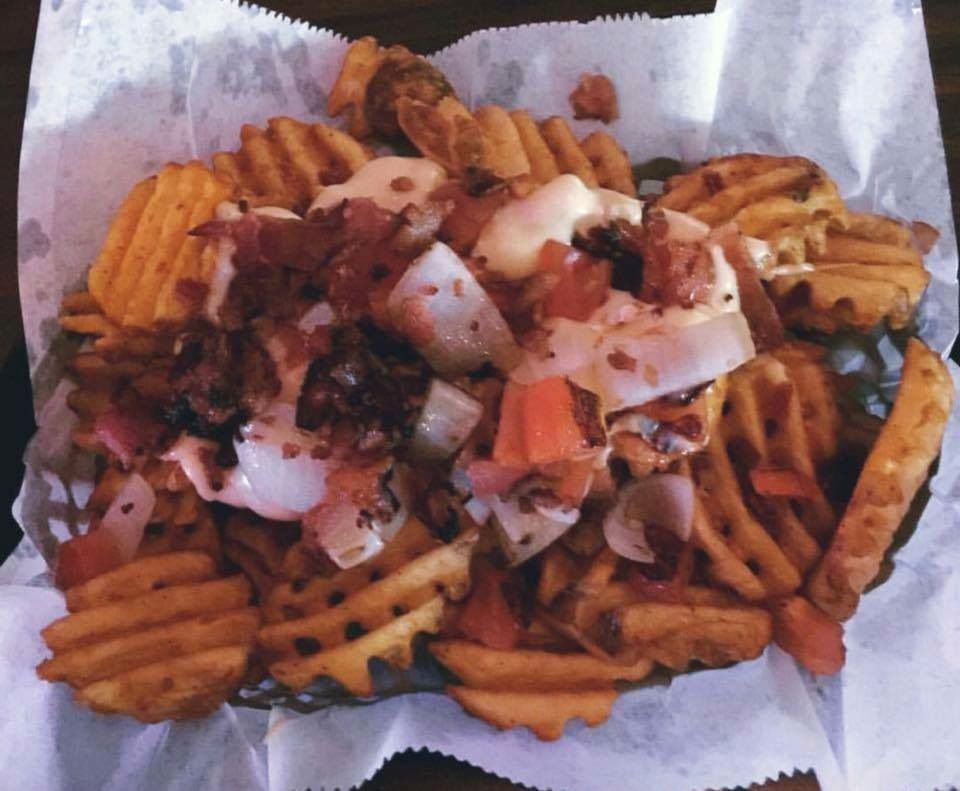 Waffle fries topped with bacon, onions, cheese, and sauce in a paper-lined basket.