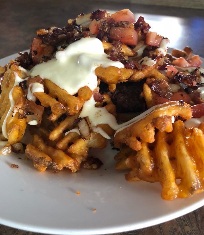 Waffle fries topped with bacon, diced tomatoes, onions, and creamy sauce on a white plate.
