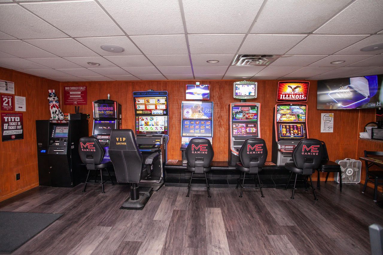 A small game room with several slot machines lined up against a wooden panel wall. Black chairs face each machine.