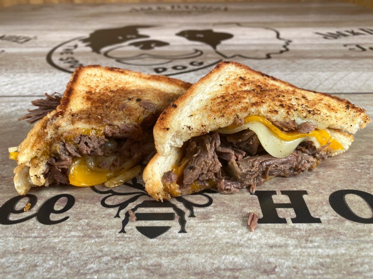 Grilled cheese sandwich with beef, onions, and two cheese types, cut in half on a wooden surface.