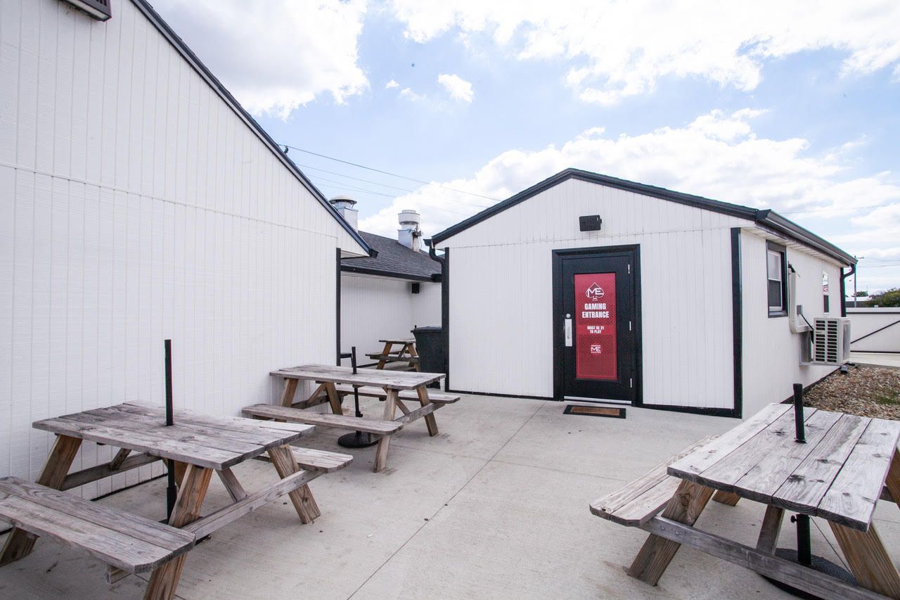 Outdoor patio with wooden picnic tables, adjacent to white buildings with black trim.