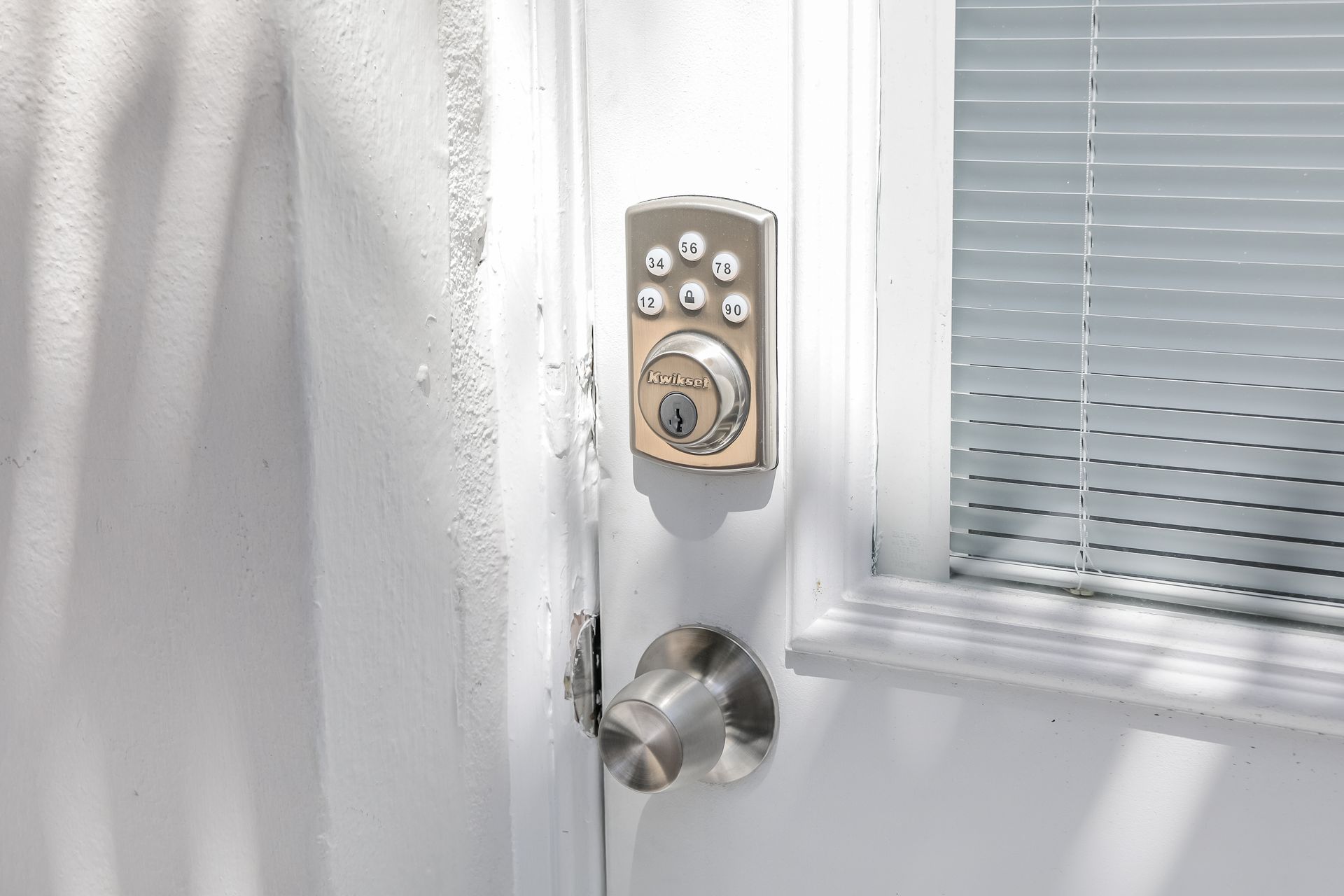 White exterior door with keypad deadbolt, doorknob, and window blinds beside it