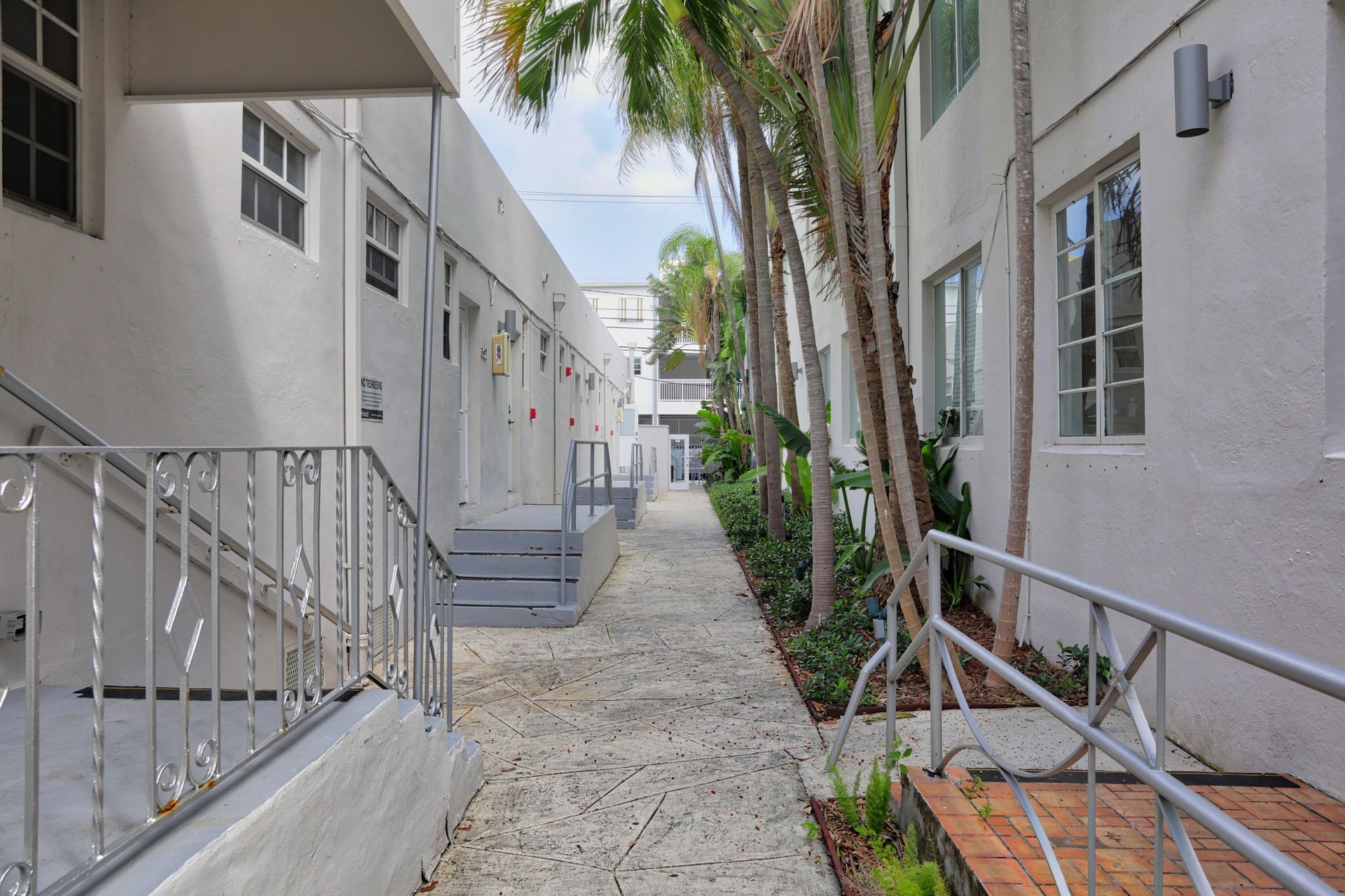 Narrow apartment walkway with stairs, white buildings, and palm trees lining the path.