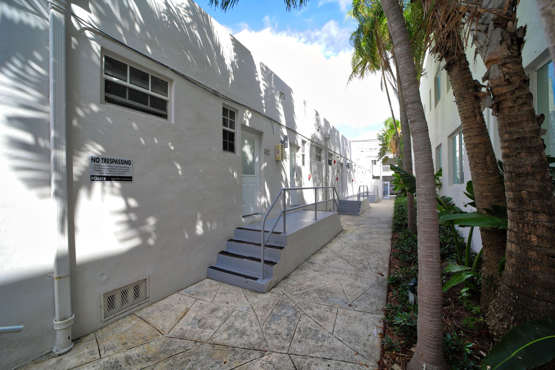 White apartment building exterior with stairs, palm trees, and a gravel walkway in bright sunlight