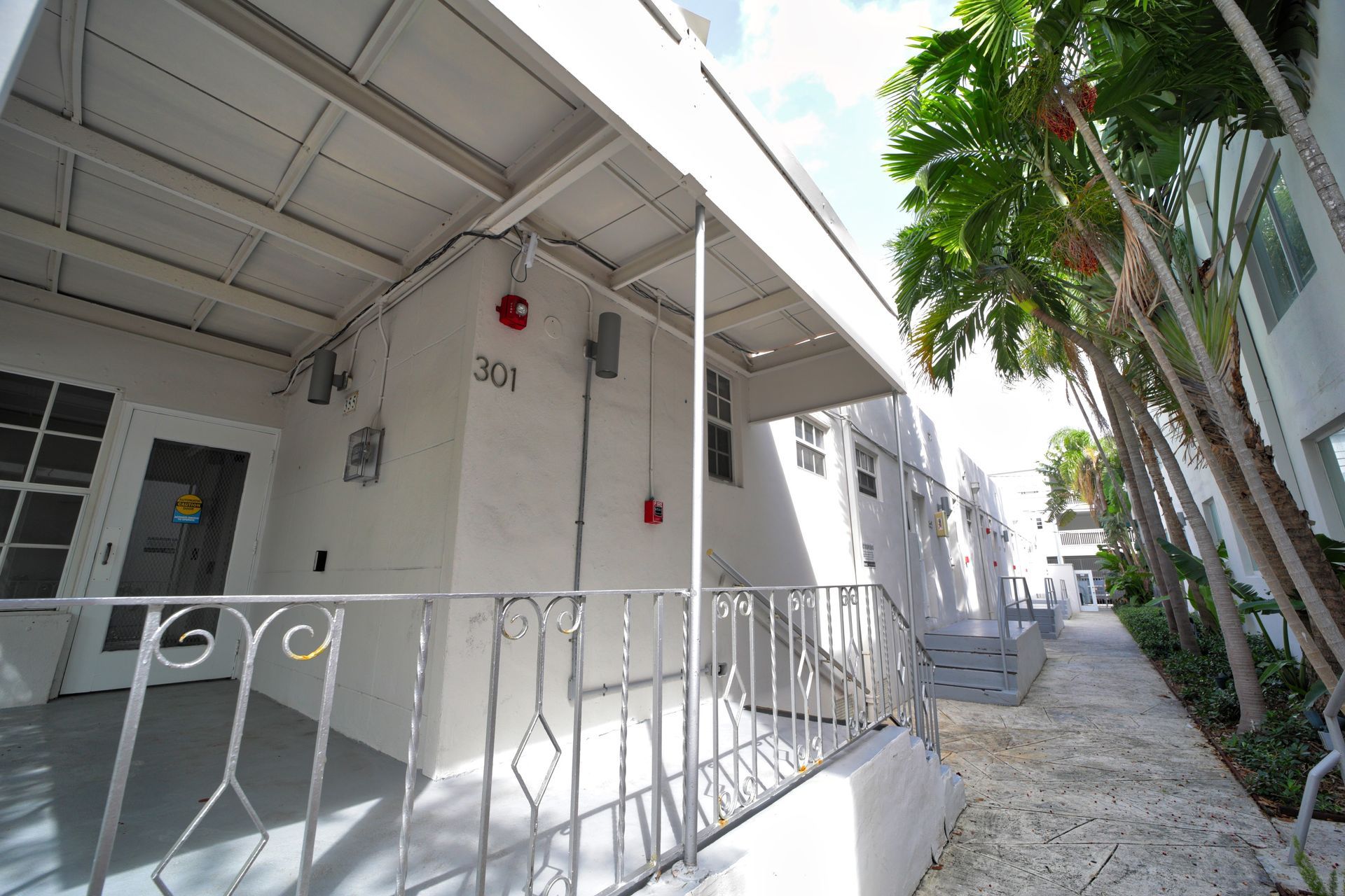 White apartment walkway with railings, red doorbell, and palm trees along a narrow side path