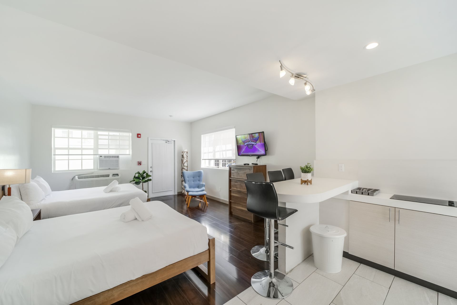 Bright modern studio with white beds, desk, dark wood floors, and wall-mounted TV