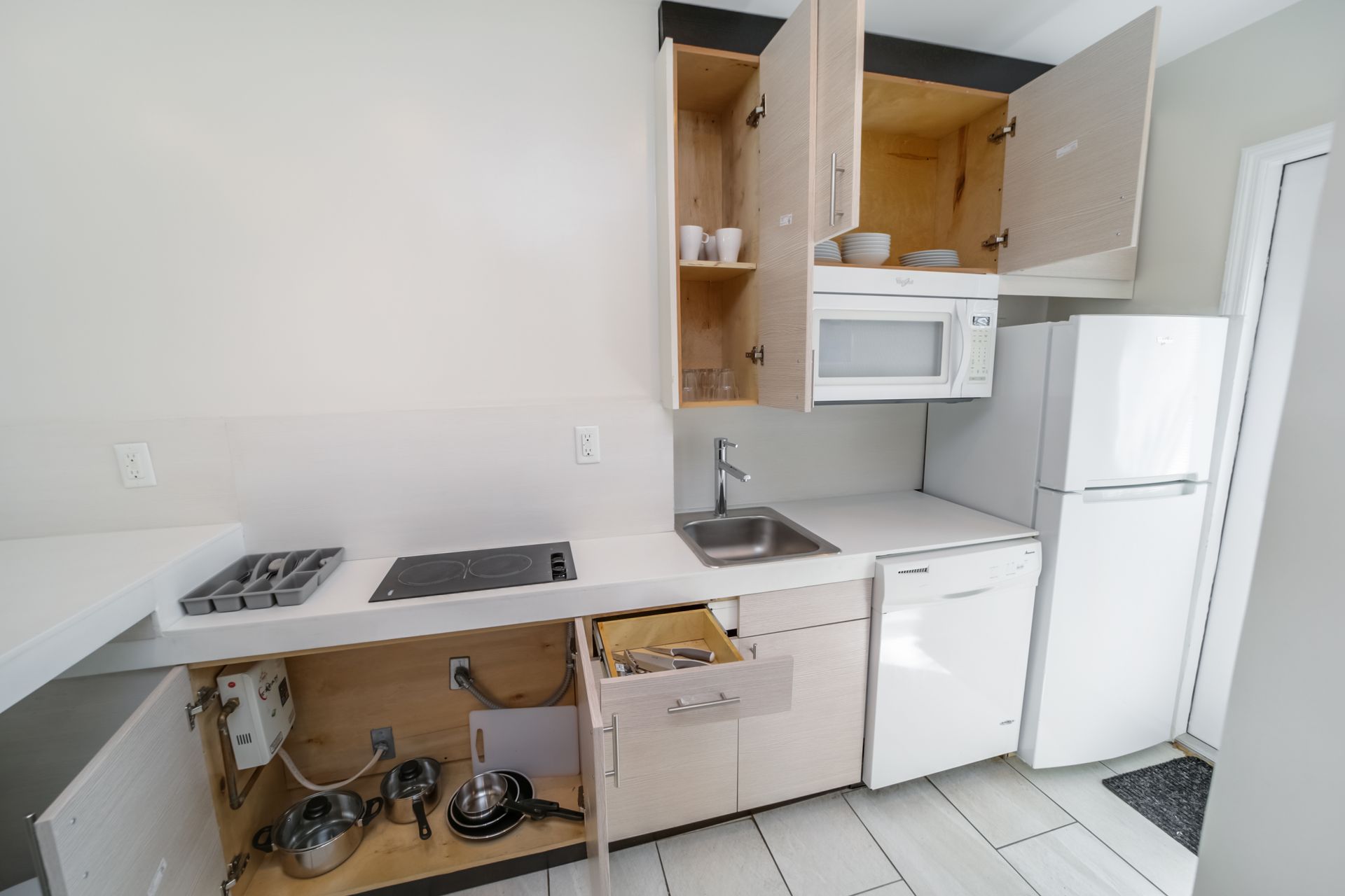 Small white kitchen with open cabinets, stove, sink, microwave, and refrigerator with doors ajar.