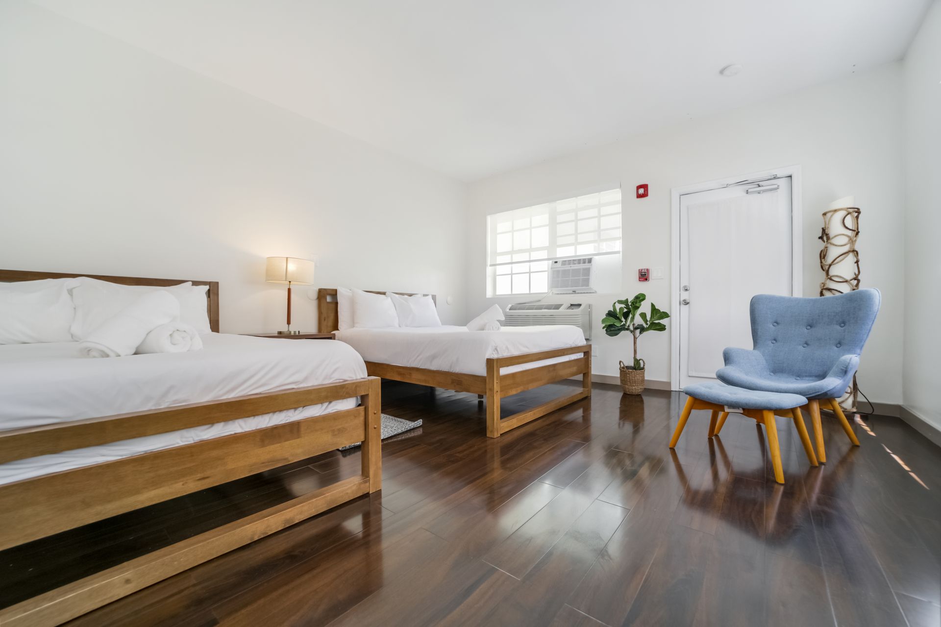 Bright bedroom with two beds, dark wood floors, blue chair, and a window with white shutters