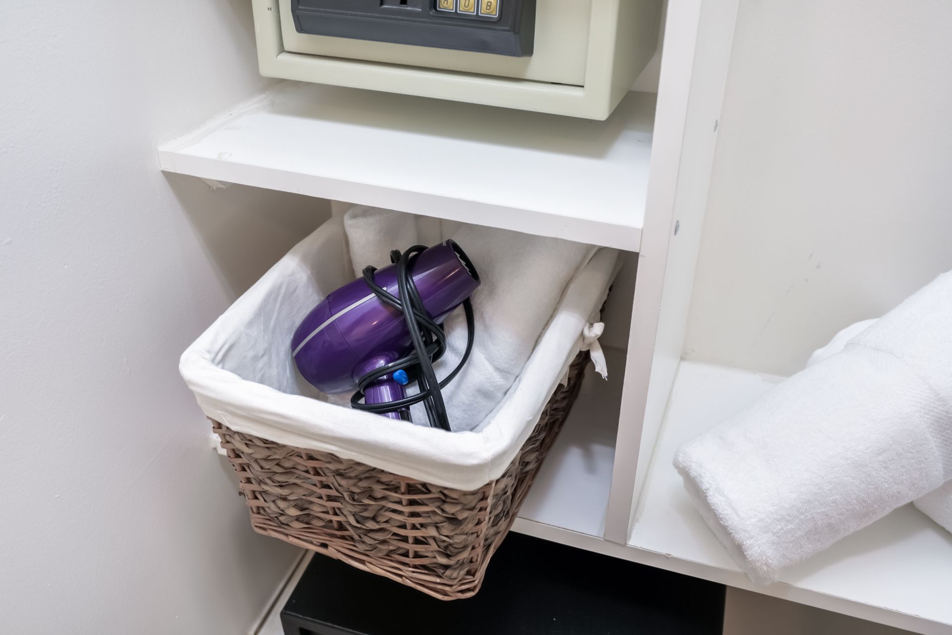 Purple hair dryer in a wicker basket on a shelf, with a rolled white towel beside it.