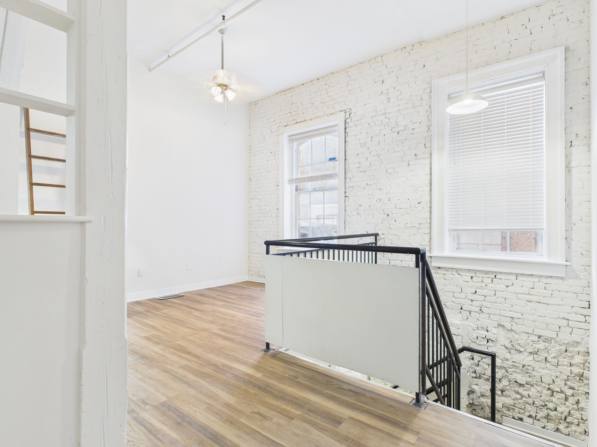Bright, empty room with wood floors, white brick walls, windows, and a staircase.