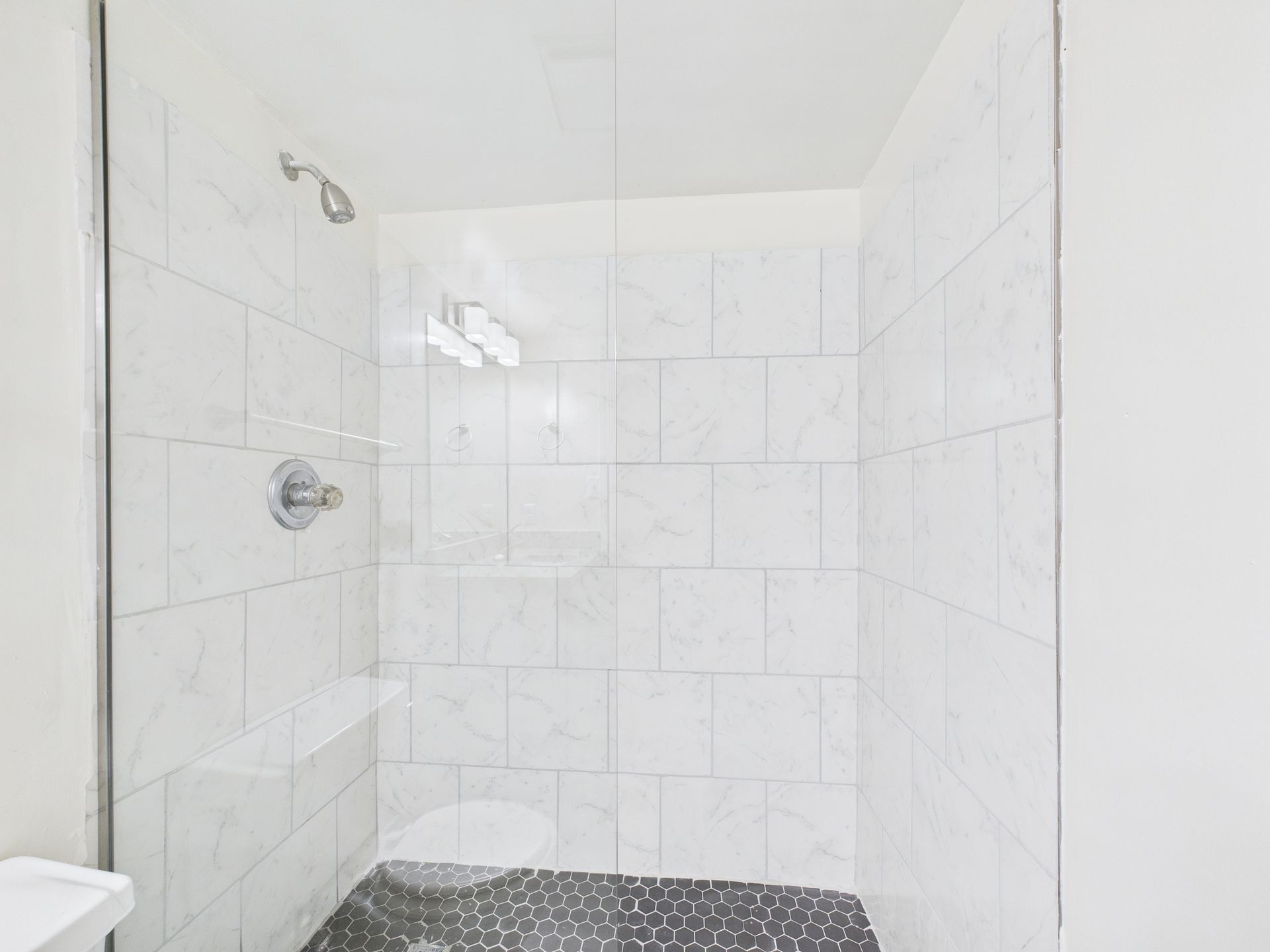 White tiled shower with black stone floor, glass door, and silver shower head and faucet.