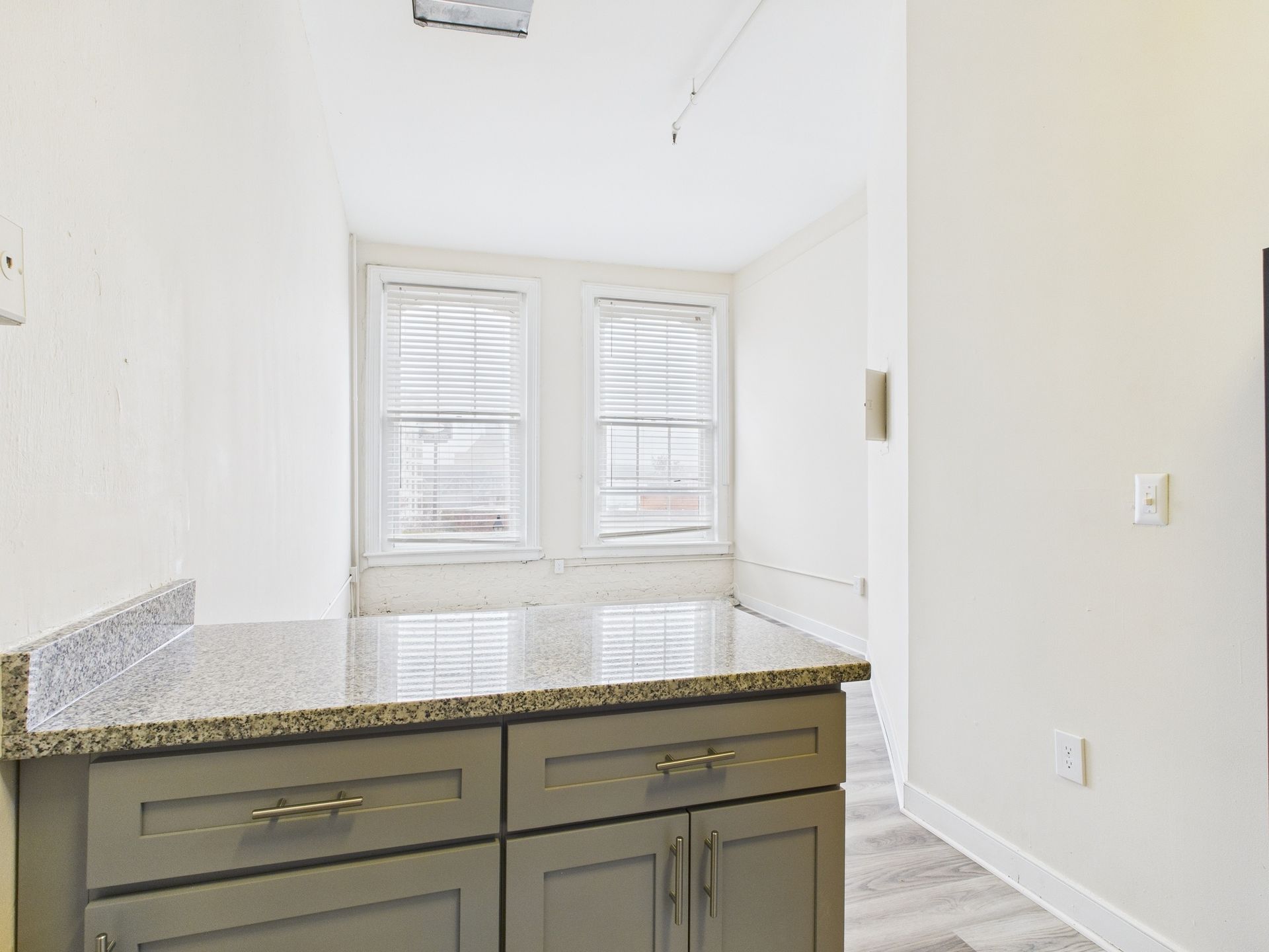 Gray cabinets with granite countertop in front of windows. White walls, grey flooring, and a hallway.