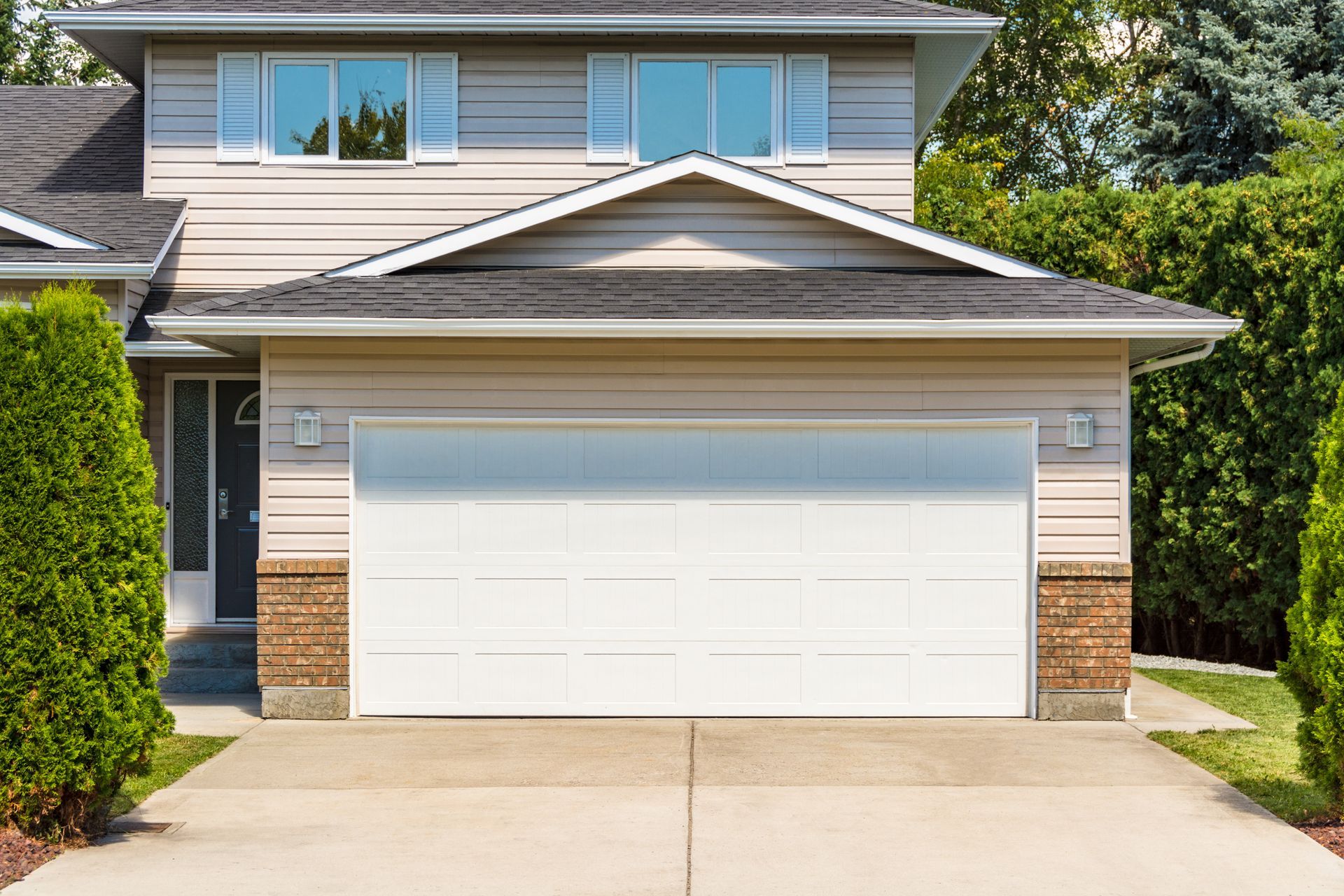 Suburban two-story house with white garage door, beige siding, and a concrete driveway in bright daylight