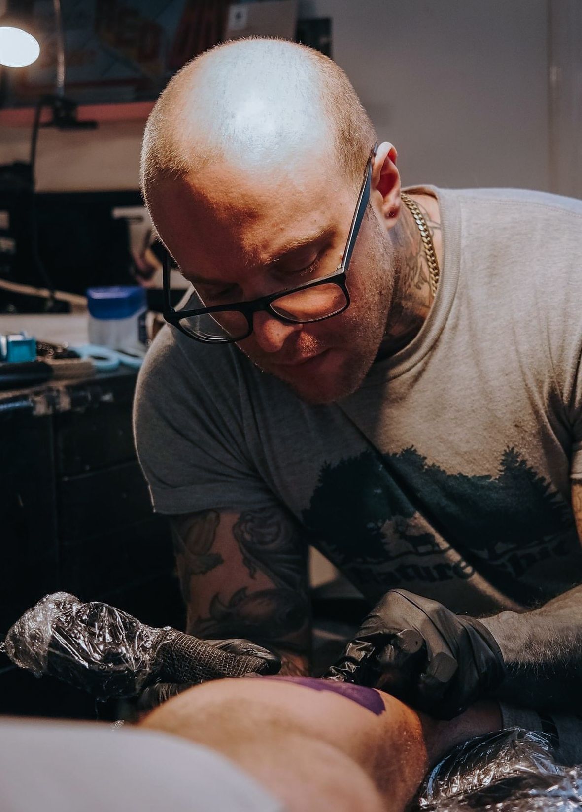 A person with tattoos on their arms stands in a studio wearing a black shirt with a 