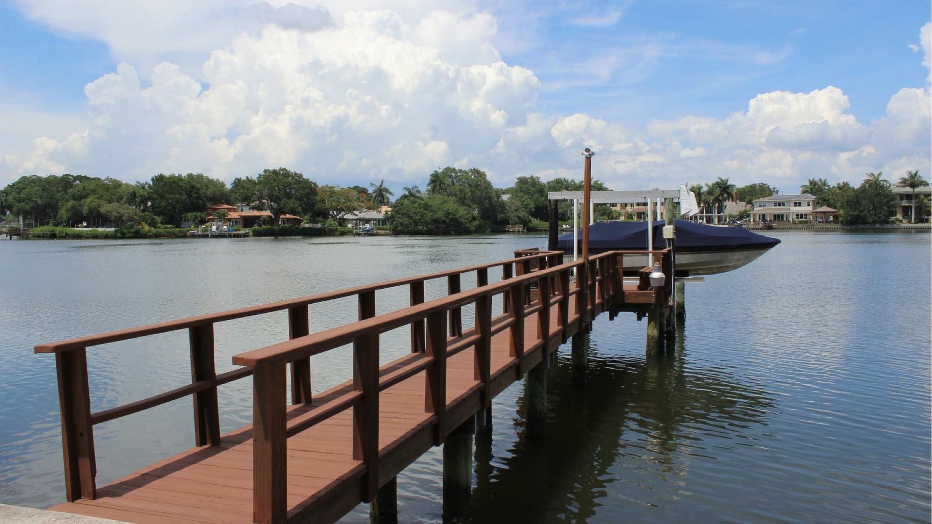 Wooden dock extends over water, boat partially covered, houses and trees in background, blue sky with clouds.