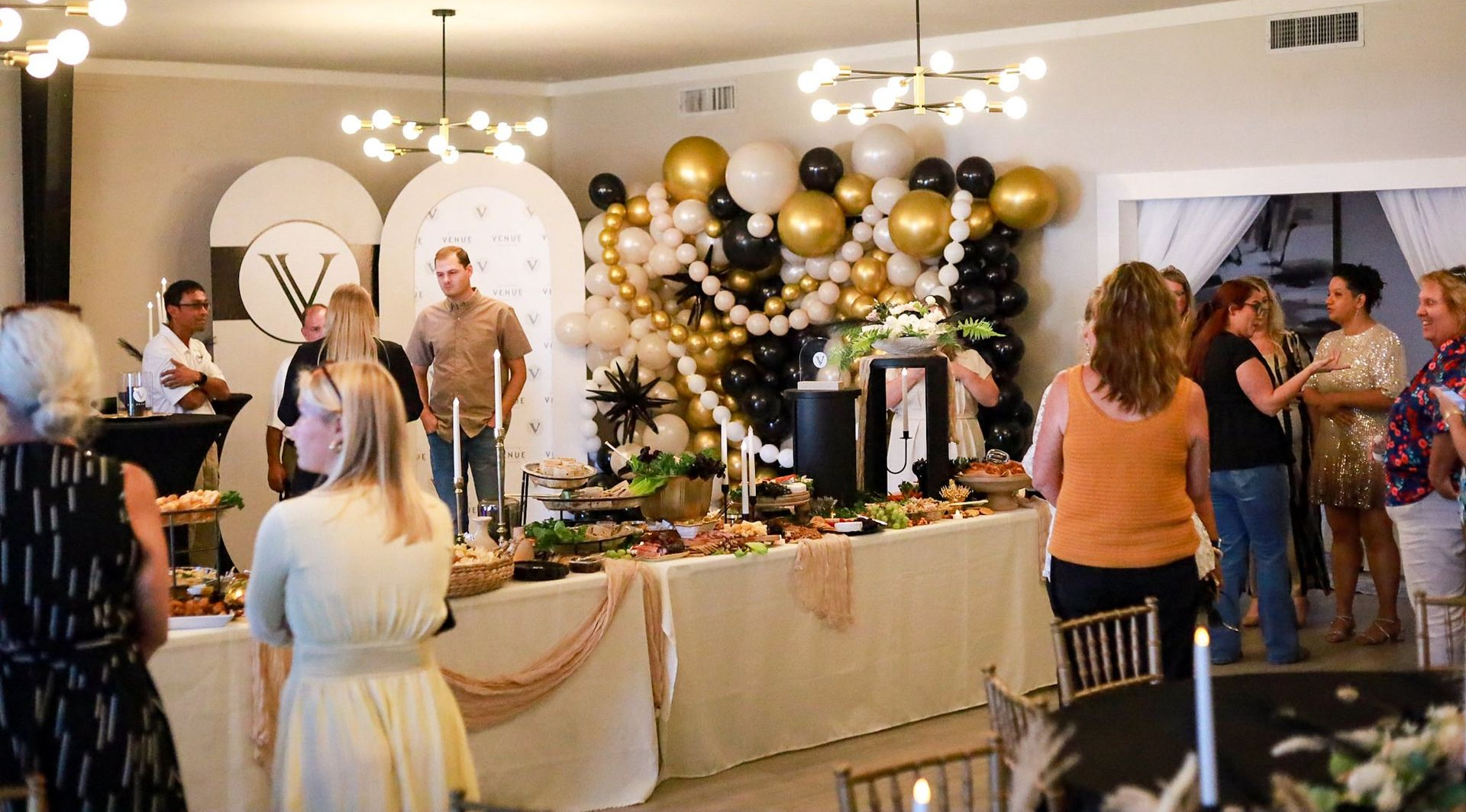 Party scene with guests, food table with gold, black, and white balloon decorations.