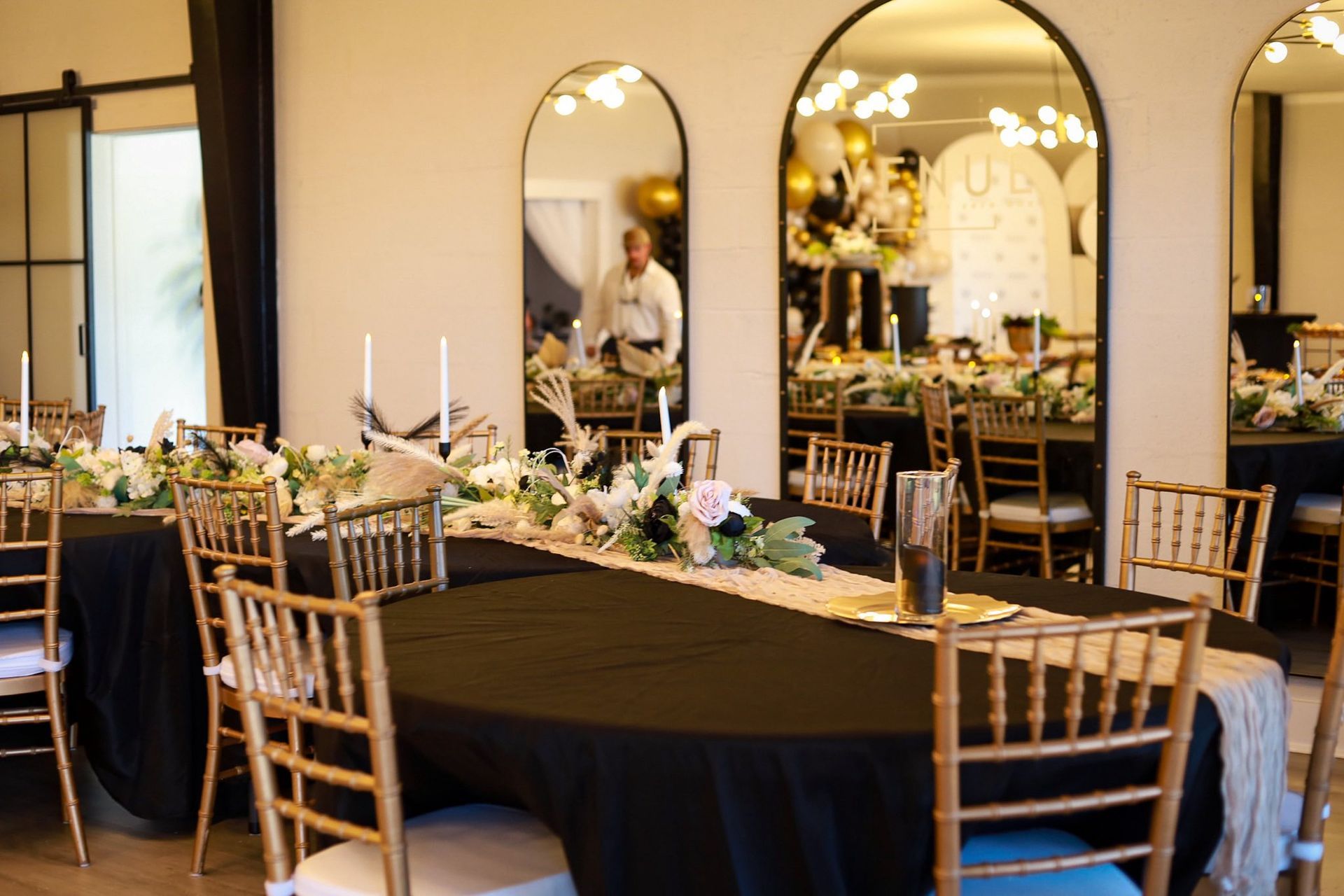 Formal dining room set for event: black tablecloth, gold chairs, mirrors, floral centerpieces.