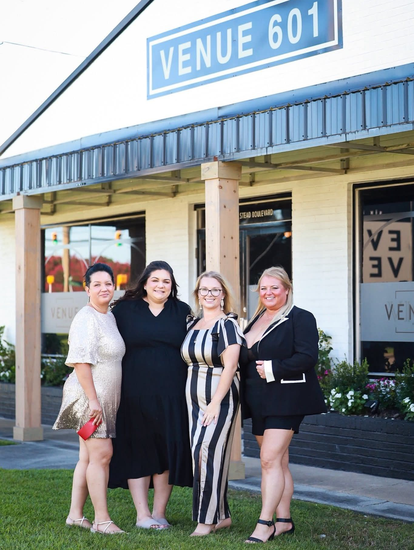 Four women posing in front of Venue 601. One in sequins, black dress, striped jumpsuit, and a suit.