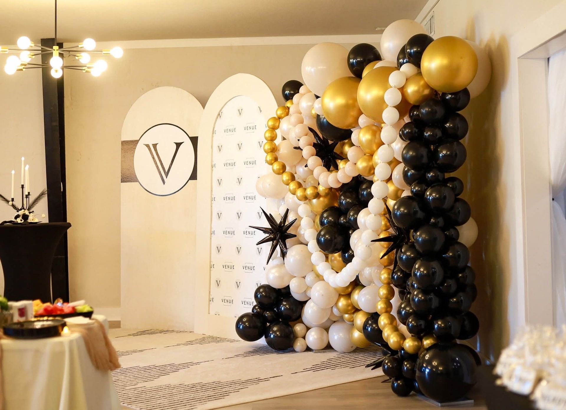 Party backdrop with balloons in black, white, and gold, and a table with food.