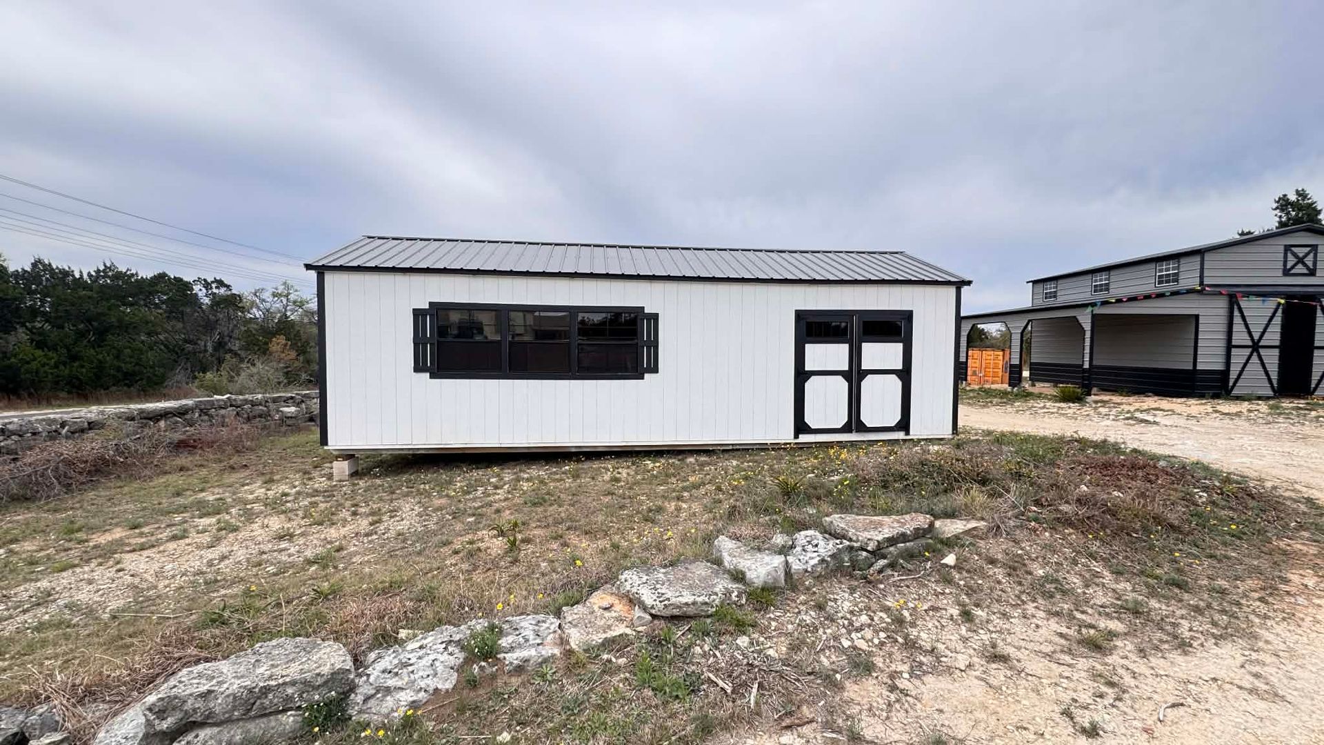 A white shed with black trim and a dark metal roof sits on a gravelly, sloped landscape under a cloudy sky.